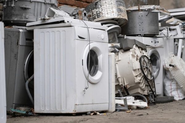 A pile of discarded washing machines and appliance parts, showcasing broken and worn-out equipment stacked outdoors.