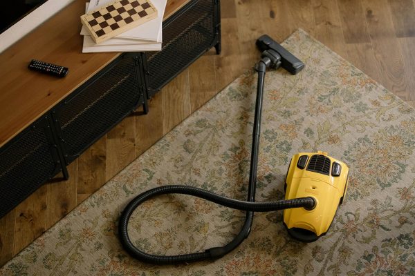 Yellow vacuum cleaner on a patterned area rug in a cozy living room with a wooden floor, media console, and remote control on top.