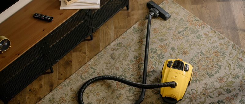 Yellow vacuum cleaner on a patterned area rug in a cozy living room with a wooden floor, media console, and remote control on top.