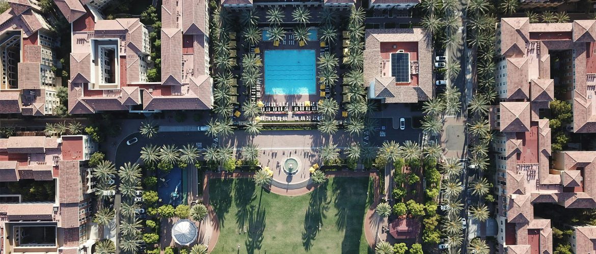 Drone view of a luxury residential complex featuring a large central pool surrounded by palm trees, well-manicured lawns, and Mediterranean-style buildings