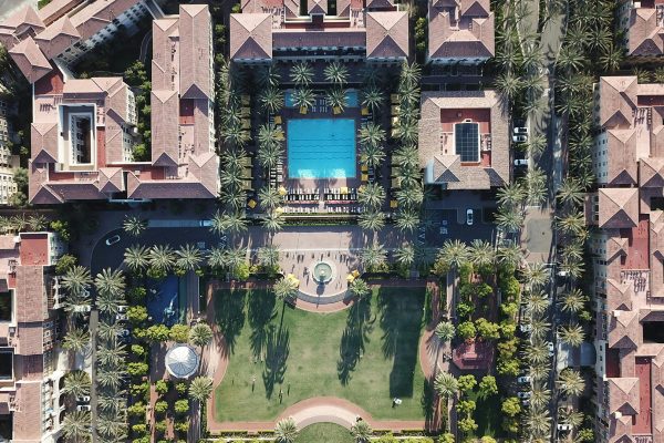 Drone view of a luxury residential complex featuring a large central pool surrounded by palm trees, well-manicured lawns, and Mediterranean-style buildings