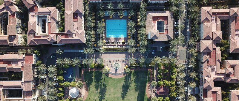 Drone view of a luxury residential complex featuring a large central pool surrounded by palm trees, well-manicured lawns, and Mediterranean-style buildings