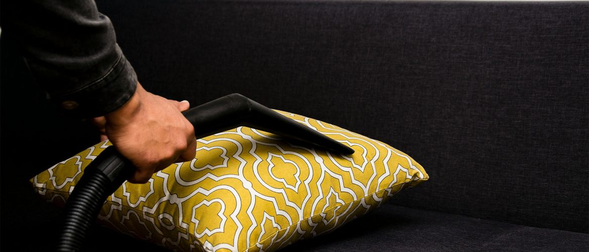 Close-up of a person using a vacuum cleaner to clean a patterned yellow pillow on a dark sofa.