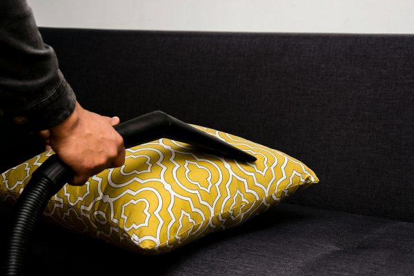 Close-up of a person using a vacuum cleaner to clean a patterned yellow pillow on a dark sofa.